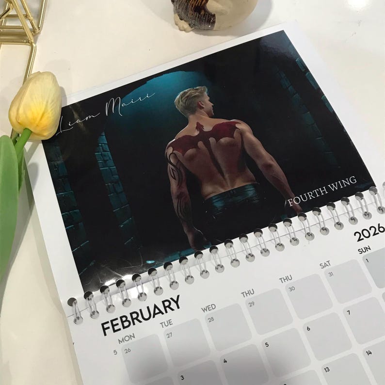May include: A spiral-bound calendar for 2026 featuring a shirtless man with a red wing tattoo on his back. The calendar shows the month of February with the text "FOURTH WING" and "Liam Maire". A yellow tulip and gold decor are in the background.