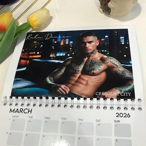 May include: A spiral-bound calendar for 2026 featuring a male model with tattoos. The calendar shows the month of March. The model is shirtless and seated. The text "Kuhn Dansan" and "CRESCENT CITY" are visible.