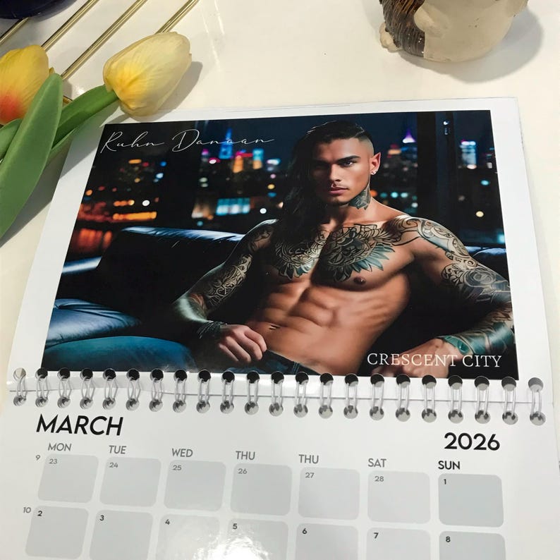 May include: A spiral-bound calendar for 2026 featuring a male model with tattoos. The calendar shows the month of March. The model is shirtless and seated. The text "Kuhn Dansan" and "CRESCENT CITY" are visible.