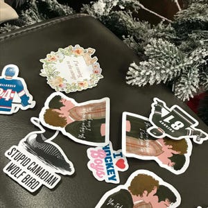 May include: A collection of stickers on a dark surface. The stickers feature various designs, including a hockey player, a floral wreath with text, a cartoon of two people kissing, and text that reads "STUPID CANADIAN WOLF BIRD" and "I HOCKEY Boys".
