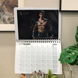 May include: A wall calendar for the year 2026, featuring a male model on the top page. The calendar shows the month of November. The text "A COURT OF THORNS AND ROSES" is visible. The calendar is white with a spiral binding.