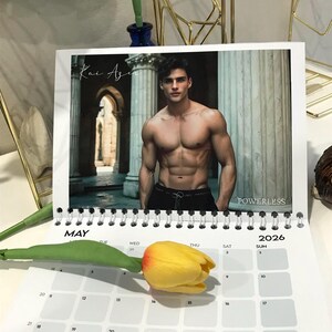 May include: A calendar for the year 2026, open to May, featuring a photograph of a shirtless man. The calendar has a spiral binding and a yellow tulip rests on the calendar page. The word "POWERLESS" is visible.