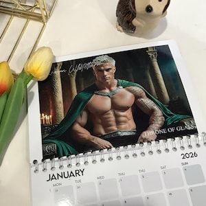 May include: A 2026 calendar featuring a fantasy illustration of a muscular person with silver hair, wearing a green cape. The calendar is open to January, with the text "Throne of Glass" visible.