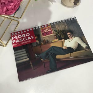 Könnte beinhalten: Ein Spiral-Kalender für 2026 mit einem Foto von Pedro Pascal. Das rote Cover trägt den weißen Schriftzug: „A Year of Pedro Pascal“. Der Kalender zeigt einen Mann in weißem Hemd und dunkler Hose auf einem gelben Sofa sitzend.