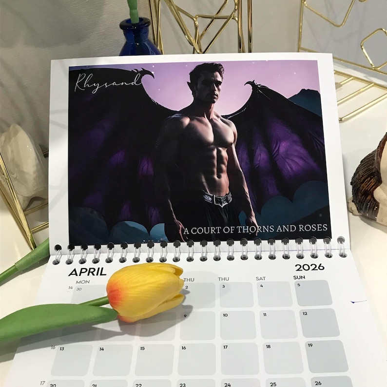 May include: A 2026 spiral-bound calendar featuring a muscular man with wings, the text "Rhysand" and "A Court of Thorns and Roses." The calendar is open to April, with a yellow tulip in the foreground.