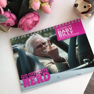 Pode incluir: Um calendário com espiral com o texto "A Year of Baby Billy 2026 Calendar" em rosa. A imagem mostra um homem de óculos de sol a fazer um gesto obsceno, com as palavras "Go Outside Nerd" em rosa.