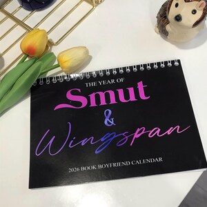 May include: A black spiral-bound calendar with the text "THE YEAR OF Smut & Wingspan" in pink and blue gradient lettering. The calendar is labeled "2026 BOOK BOYFRIEND CALENDAR". Decorative tulips and a hedgehog figurine are also visible.