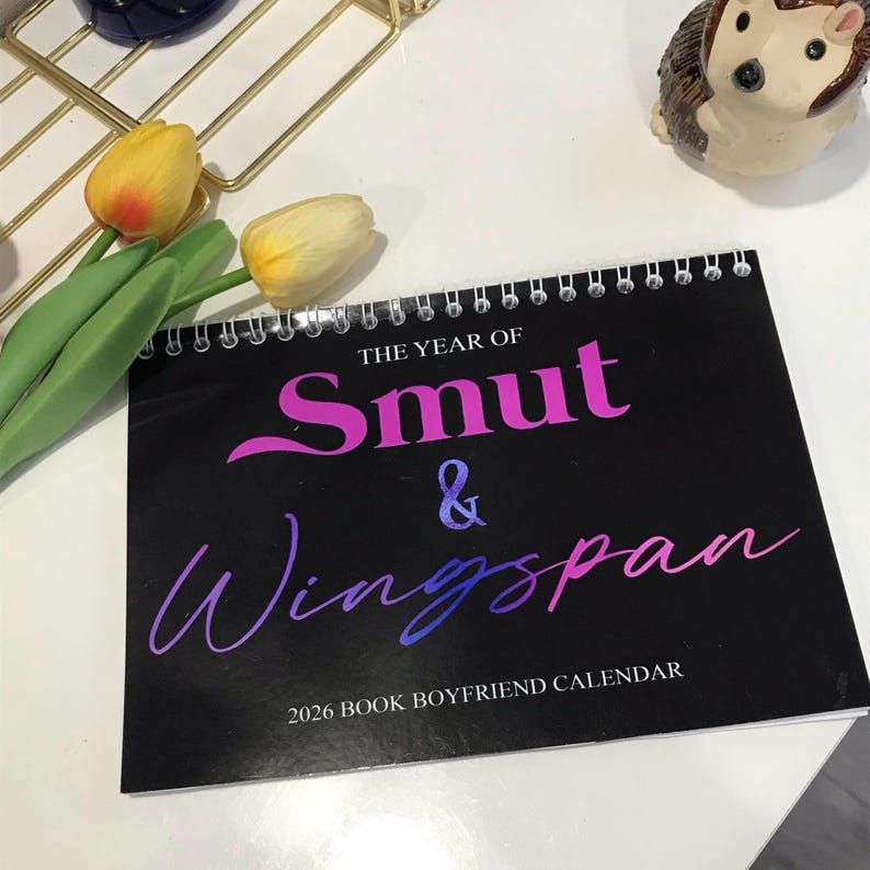 May include: A black spiral-bound calendar with the text "THE YEAR OF Smut & Wingspan" in pink and blue gradient lettering. The calendar is labeled "2026 BOOK BOYFRIEND CALENDAR". Decorative tulips and a hedgehog figurine are also visible.