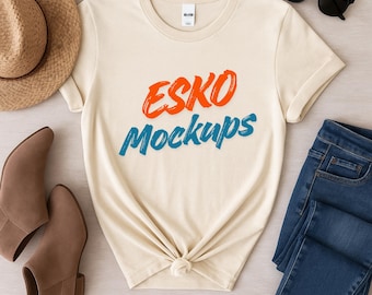 Op de afbeelding: Crèmekleurig T-shirt met de tekst "ESKO Mockups" in oranje en blauw. Het shirt is gestyled met een blauwe jeans, bruine laarzen, een strohoed en een zwarte zonnebril. Het shirt is in de taille geknoopt.