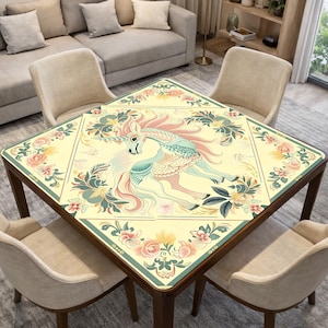 May include: A square wooden table with a cream-colored top featuring a unicorn design. The unicorn is depicted in pastel colors, surrounded by floral patterns. The table is surrounded by four beige chairs.