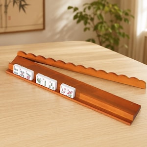 May include: A wooden mahjong tile rack with a scalloped edge, holding several white tiles with colorful illustrations. The rack is made of dark brown wood and sits on a light-colored wooden surface. The tiles feature floral and other decorative designs.