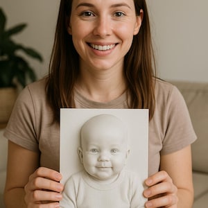 Puede incluir: Una mujer sostiene un retrato en bajorrelieve enmarcado de un bebé. El bebé está representado con una camiseta de manga larga acanalada de color claro. El retrato es una escultura tridimensional con un acabado liso y mate.