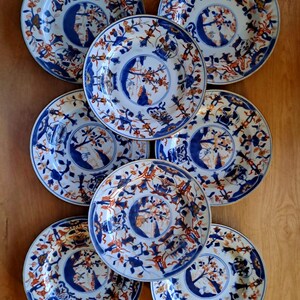 Op de afbeelding: Een verzameling van acht antieke keramische borden. Elk bord heeft een centraal blauw ontwerp, omgeven door ingewikkelde patronen in blauw en oranje tegen een witte achtergrond. De borden zijn gerangschikt op een houten oppervlak.