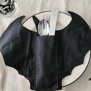 May include: Black bat-shaped fabric place card holder with a silver fork, knife, and spoon inserted into the pocket. The holder is on a white plate.