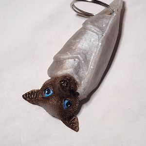 Può includere: Un portachiavi a forma di pipistrello marrone e bianco con occhi blu. Il pipistrello è arrotolato in una posizione di sonno con le ali avvolte attorno al suo corpo. Il portachiavi è fatto di un materiale morbido e flessibile.