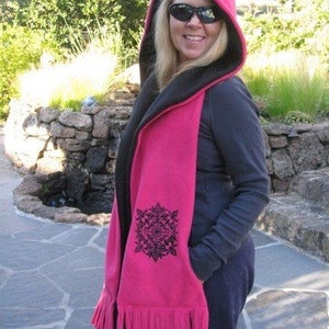 May include: A pink hooded scarf with a black interior and a decorative black floral design. The scarf has a fringe at the bottom and is made of a soft material. The hood is pointed at the top.