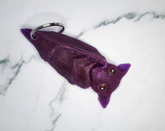 May include: Purple bat-shaped keychain with a silver ring. The bat has large, black eyes and is curled up in a sleeping position.