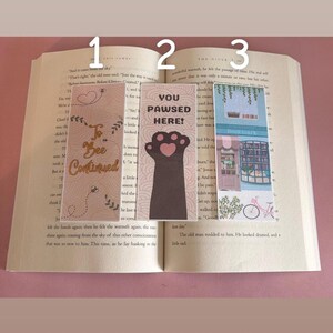 May include: Three rectangular bookmarks placed on an open book. The first is pink with bee illustrations and the text To Bee Continued. The second is white with a black paw print and the text YOU PAWSED HERE!. The third is blue with a bookstore illustration.