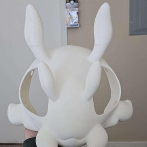 Op de afbeelding: Een witte, onbeschilderde masker met grote, rechtopstaande oren en afgeronde kenmerken. Het masker heeft oogopeningen en een gebogen vorm, vastgehouden door een persoon. Het masker lijkt een cosplay- of kostuumstuk te zijn.