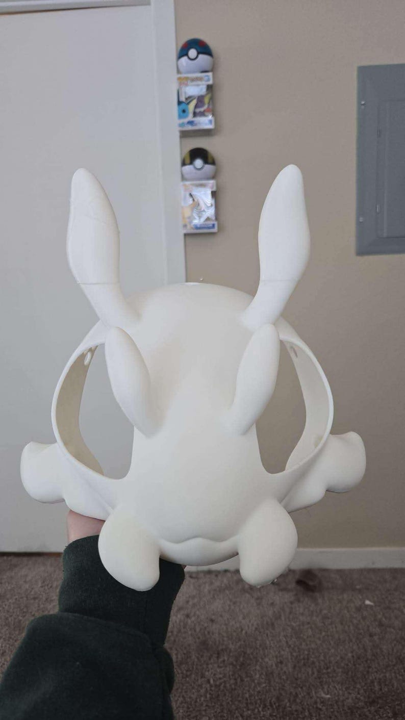 Op de afbeelding: Een witte, onbeschilderde masker met grote, rechtopstaande oren en afgeronde kenmerken. Het masker heeft oogopeningen en een gebogen vorm, vastgehouden door een persoon. Het masker lijkt een cosplay- of kostuumstuk te zijn.