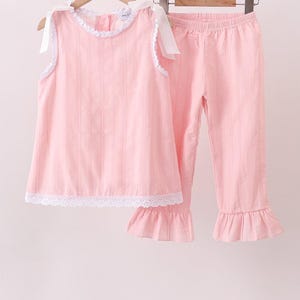 May include: A light pink sleeveless top and matching pants set. The top features white ribbon shoulder ties, a round neckline with white lace trim, and a white lace hem. The pants have an elastic waistband and ruffled hems at the bottom of each leg.