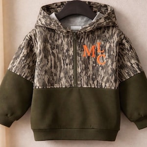 May include: A long-sleeve hooded sweatshirt featuring a half-zip closure. The upper portion and hood display a brown and green tree bark camouflage pattern, while the lower section and sleeves are solid olive green. The letters MLC are in orange.
