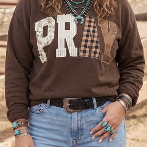 Op de afbeelding: Een bruin sweatshirt met lange mouwen en het woord PRAY in grote, verschillend bedrukte stoffen letters op de borst. De letters hebben bloemmotieven, strepen, ruitjes en effen bruine texturen. Het sweatshirt wordt gedragen met een lichtblauwe jeans.