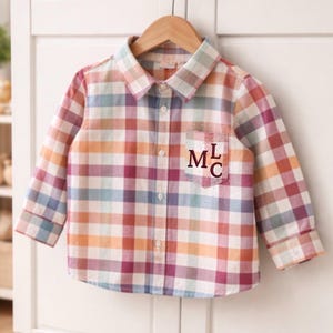 May include: A long-sleeve button-down shirt featuring a multi-colored plaid pattern in shades of pink, orange, blue, and white. The shirt has a pointed collar, a front button placket, and a chest pocket embroidered with the dark red letters MLC.