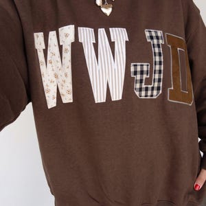 Op de afbeelding: Een donkerbruine sweater met ronde hals en de letters WWJD op de borst geappliqueerd. Elke letter is gemaakt van verschillende stoffen: bloemenprint, verticale strepen, zwart-wit gingham ruitje en een effen bruine suède-achtige stof.