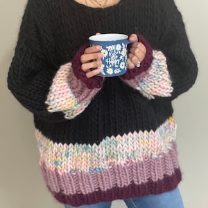 May include: A black chunky knit jumper with colourful stripes on the sleeves and bottom hem. The jumper is oversized and has a relaxed fit. The model is holding a blue mug with the words "Be Happy" on it.