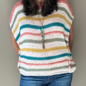 Op de afbeelding: Een gestreepte gebreide top in wit, roze, geel, turquoise en groen. De top heeft een ronde hals en korte mouwen. De zijnaden zijn gedetailleerd met knopen. Een lange turquoise en gouden ketting hangt van de halslijn. Het model draagt een spijkerbroekshort.