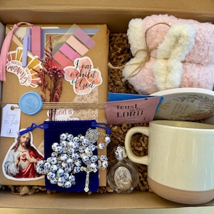 May include: A gift box filled with religious items. Includes a white and pink mug, a rosary, a small perfume bottle, a pink plush item, and a small dish. The box also contains stickers, a bookmark, and a card with the text "Trust in the Lord".
