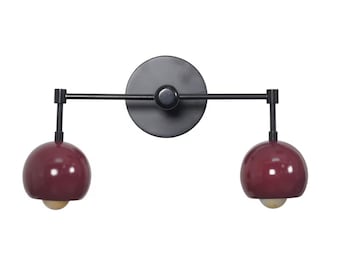 May include: A modern wall sconce with a matte black finish. It features a circular backplate, a horizontal arm, and two burgundy-colored, spherical light fixtures. The sconce is designed for indoor use and provides ambient lighting.