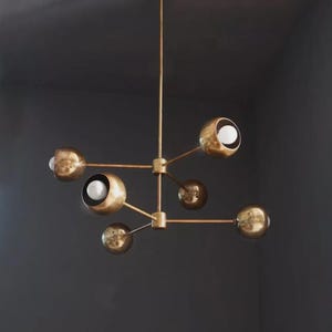 May include: A modern brass chandelier with six spherical light fixtures. Each sphere has a partially open design, revealing a bright white light bulb. The fixture is suspended from a single rod.