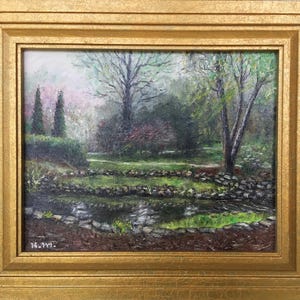 May include: Oil painting of a serene garden scene in a gold frame. The artwork depicts a pond with stone borders, lush green grass, and trees with vibrant foliage. The painting has a soft, impressionistic style, with a focus on natural light and color.