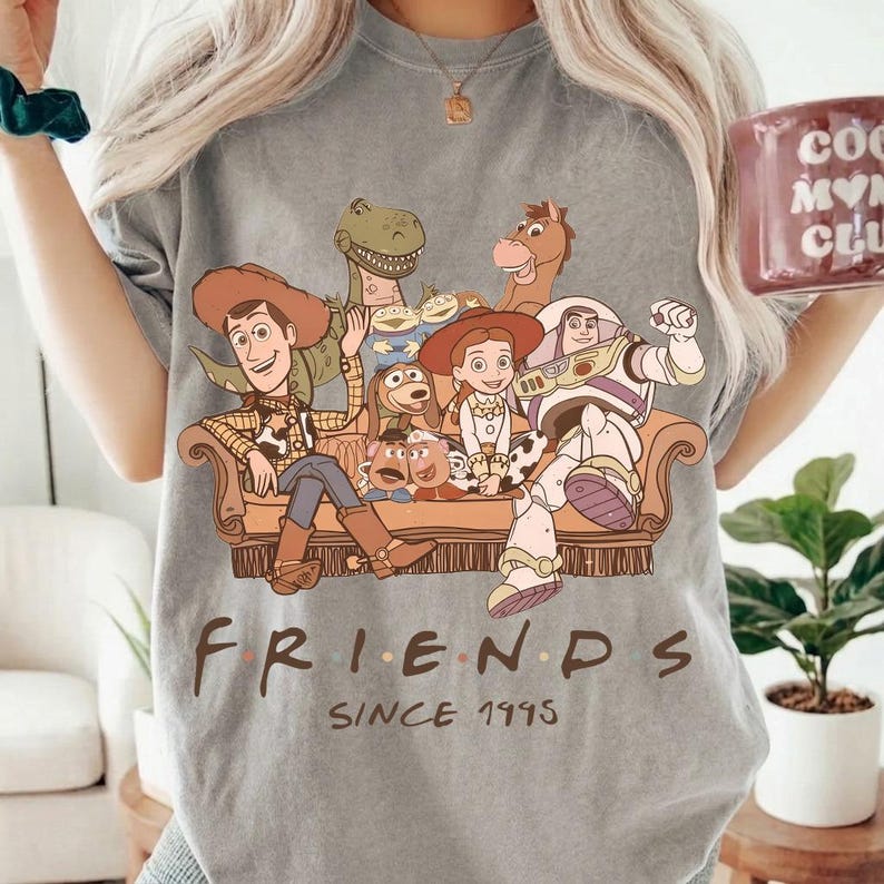 May include: Gray t-shirt featuring a graphic of Toy Story characters on a brown couch. The word "FRIENDS" is printed below the graphic, with "SINCE 1995" underneath. The shirt has a crew neck and short sleeves.