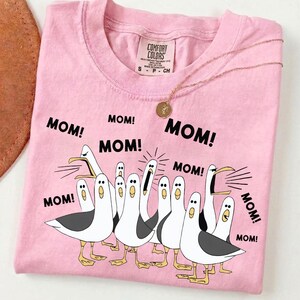 Pu&ograve; includere: Maglietta rosa Comfort Colors con una grafica di gabbiani che urlano "MOM!" in testo nero. Una collana d'oro con un ciondolo &egrave; appoggiata sulla maglietta. La maglietta ha un girocollo e maniche corte.