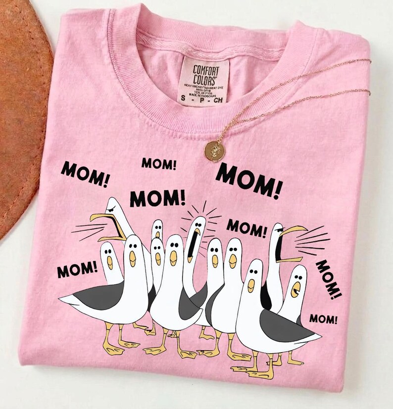 Pu&ograve; includere: Maglietta rosa Comfort Colors con una grafica di gabbiani che urlano "MOM!" in testo nero. Una collana d'oro con un ciondolo &egrave; appoggiata sulla maglietta. La maglietta ha un girocollo e maniche corte.