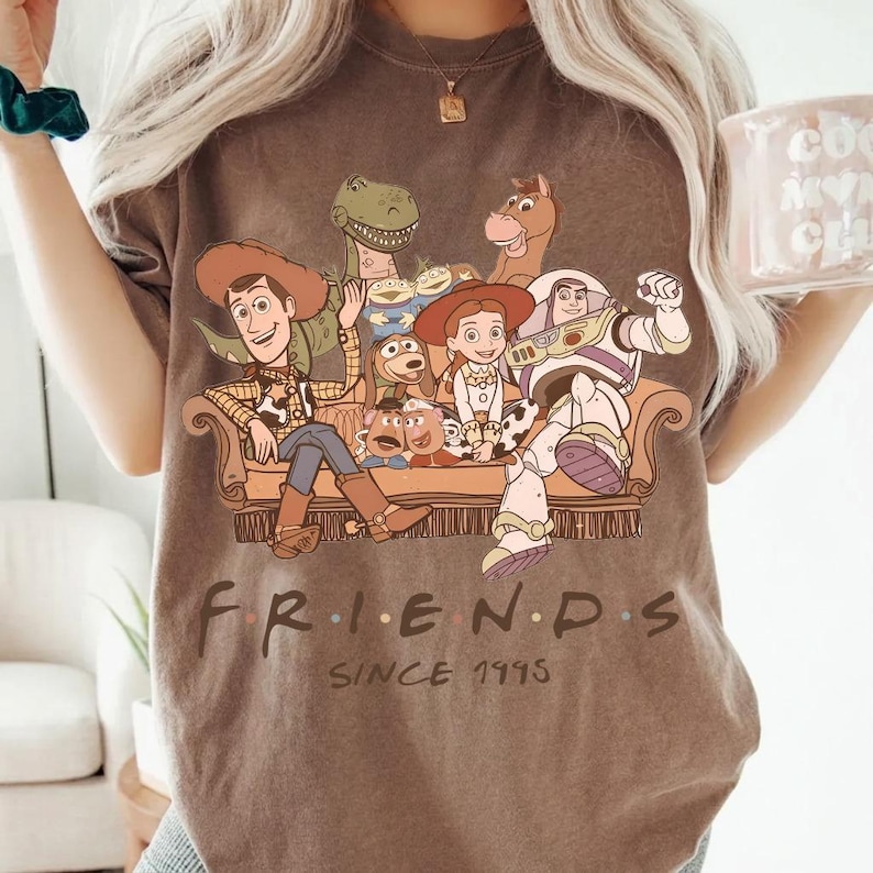 May include: Brown t-shirt featuring a graphic of characters from the movie Toy Story, including Woody, Buzz Lightyear, and others, seated on a couch. The text "FRIENDS SINCE 1995" is printed below the image. The shirt has a vintage, worn look.