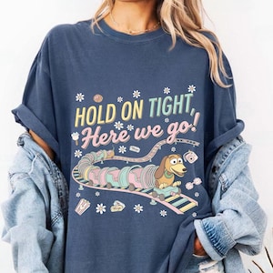 Może przedstawiać: Granatowa koszulka z napisem "HOLD ON TIGHT, Here we go!" oraz kreskówkową ilustracją jamnika na kolejce górskiej. Projekt zawiera pastelowe kolory i kwiatowe akcenty.