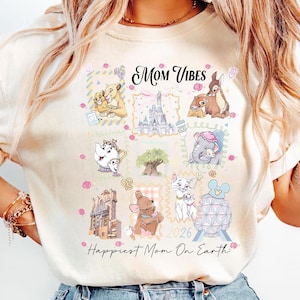 May include: Cream-colored t-shirt with the words "Mom Vibes" and "Happiest Mom On Earth." The shirt features cartoon images of Disney characters in postage stamp-like squares, including Simba, Bambi, and Marie. The year "2026" is also visible.