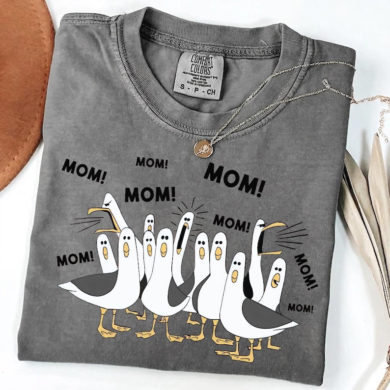 Pu&ograve; includere: T-shirt grigio Comfort Colors con una grafica di gabbiani e la parola "MOM!" ripetuta. I gabbiani sono bianchi con ali grigie e zampe gialle. Una collana d'argento con un ciondolo &egrave; drappeggiata sulla maglietta.
