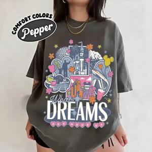 Puede incluir: Camiseta color pimienta con un dise&ntilde;o gr&aacute;fico colorido que presenta un castillo, una nave espacial y otras ilustraciones. El texto "What Dreams Are Made Of" se muestra en una fuente estilizada. La camiseta tambi&eacute;n tiene el texto "Comfort Colors" y "Pepper".