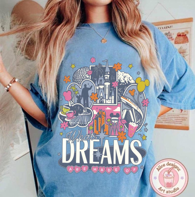 Puede incluir: Camiseta azul claro con un dise&ntilde;o gr&aacute;fico colorido que presenta un castillo, fuegos artificiales y elementos de parque tem&aacute;tico. El texto "What Dreams Are Made Of" se muestra de forma destacada.