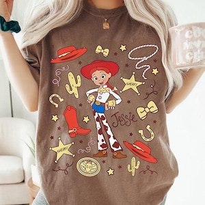 Puede incluir: Camiseta marr&oacute;n con una ilustraci&oacute;n de dibujos animados de Jessie de Toy Story. El dise&ntilde;o incluye un sombrero rojo, botas, insignias de sheriff y el nombre "Jessie". Elementos adicionales incluyen un cactus, una herradura y una cuerda.