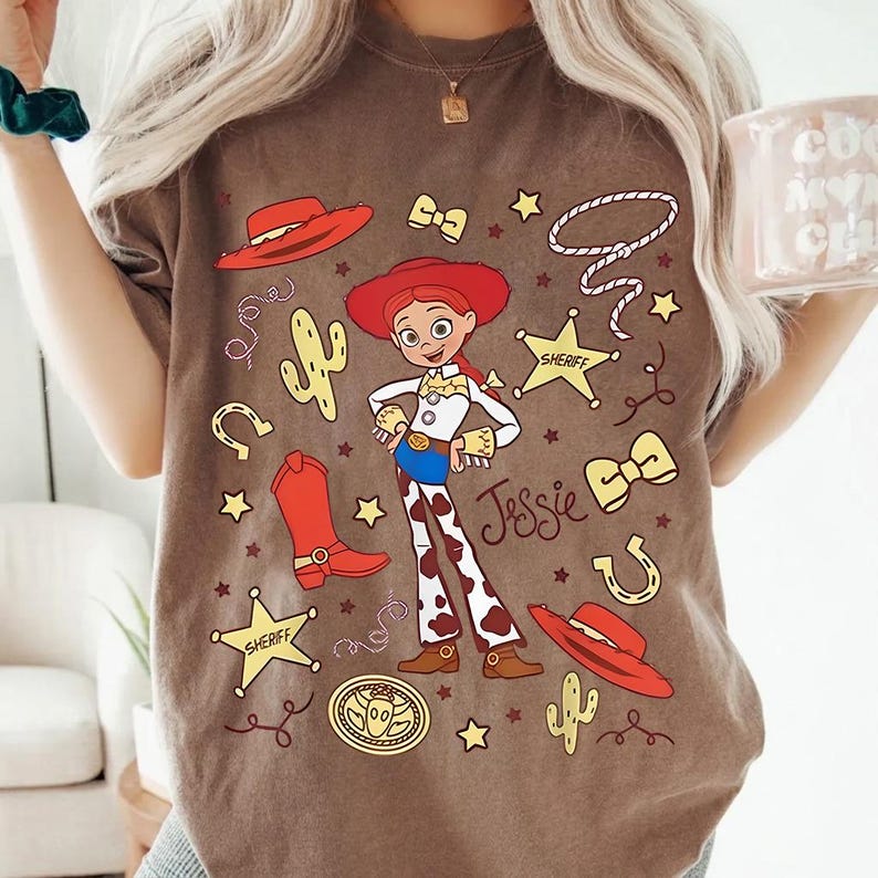 Puede incluir: Camiseta marr&oacute;n con una ilustraci&oacute;n de dibujos animados de Jessie de Toy Story. El dise&ntilde;o incluye un sombrero rojo, botas, insignias de sheriff y el nombre "Jessie". Elementos adicionales incluyen un cactus, una herradura y una cuerda.