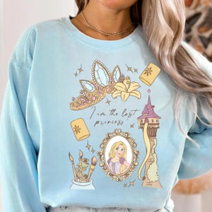May include: Pale blue sweatshirt with a graphic of a princess crown, a lily, floating lanterns, and a tower. The text "I am the lost princess" is written below the crown. A framed portrait of a blonde princess is also included.
