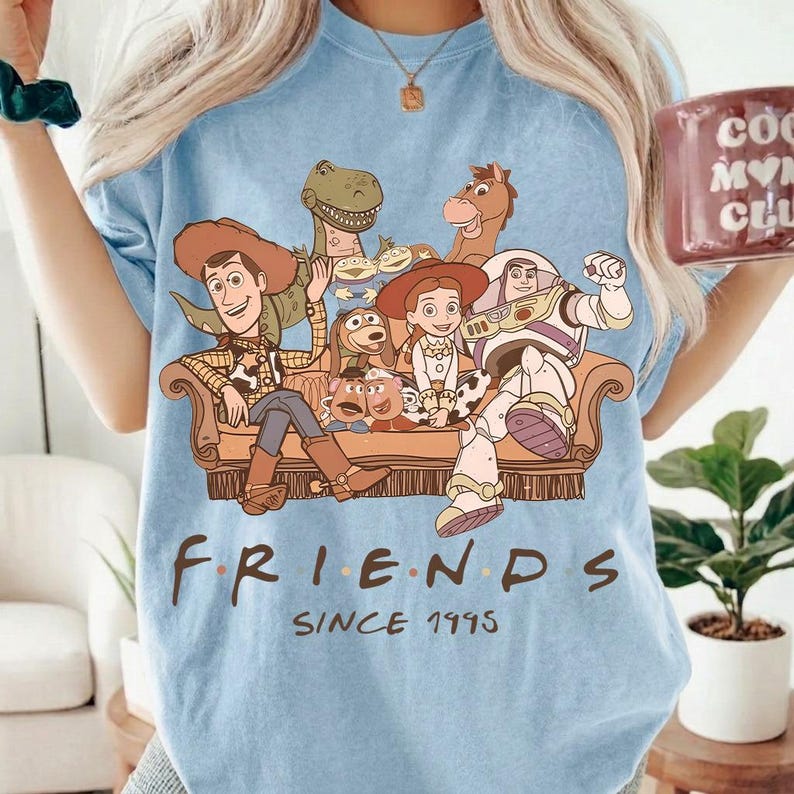 May include: Light blue t-shirt featuring a graphic of Toy Story characters on a brown couch. The word "FRIENDS" is printed below the graphic, with "SINCE 1995" underneath. The shirt is a light blue color.