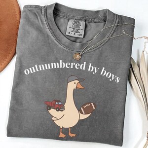 May include: Gray t-shirt with the phrase "outnumbered by boys" in white. A cartoon goose wearing a baseball cap and holding a football and a red truck is printed on the front. A silver necklace with a pendant is on the shirt.