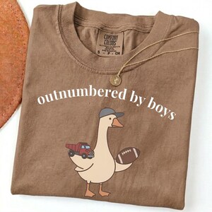 May include: A brown Comfort Colors t-shirt with the phrase "outnumbered by boys" in white. The shirt features a cartoon goose wearing a baseball cap and holding a football and a red truck. A gold necklace is draped across the shirt.